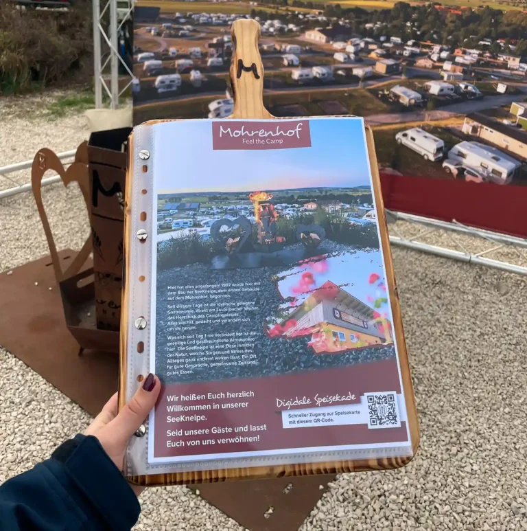 Eine Hand hält eine gedruckte Speisekarte auf einem Holzbrett vor dem Mohrenhof Franken