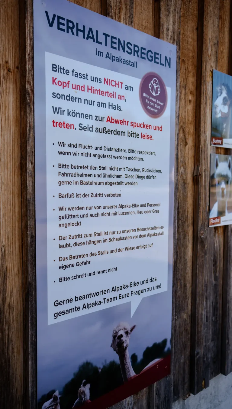 Großes Schild mit Verhaltensregeln im Alpakastall auf Deutsch, befestigt an einer Holzwand.