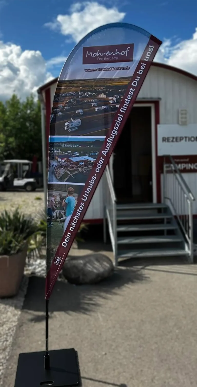 Gedruckte Drop-Flag-Fahne des Campingplatzes Mohrenhof vor der Rezeption aufgestellt