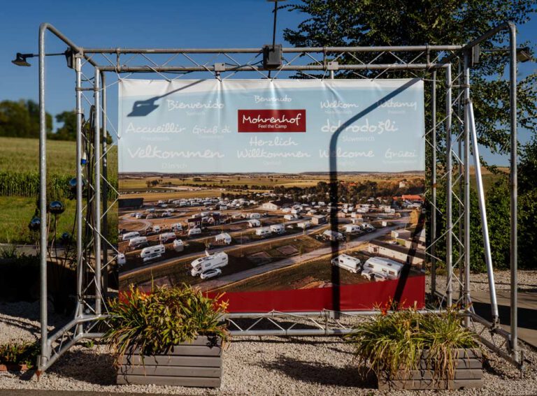 Willkommensbanner des Campingplatzes Mohrenhof auf Metallgestell mit Luftaufnahme des Geländes und Grußformeln