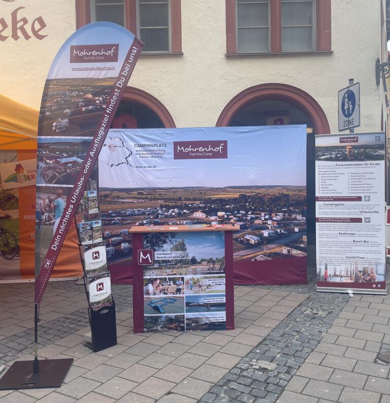 Infostand des Campingplatzes Mohrenhof im Freien mit Werbewand, Theke, Roll-up und Drop-Flag-Fahne