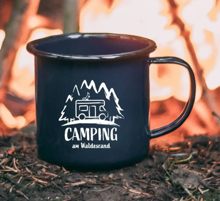 Blaue Emaille-Tasse mit weißem Logo Camping am Waldesrand vor einem gemütlichen Lagerfeuer
